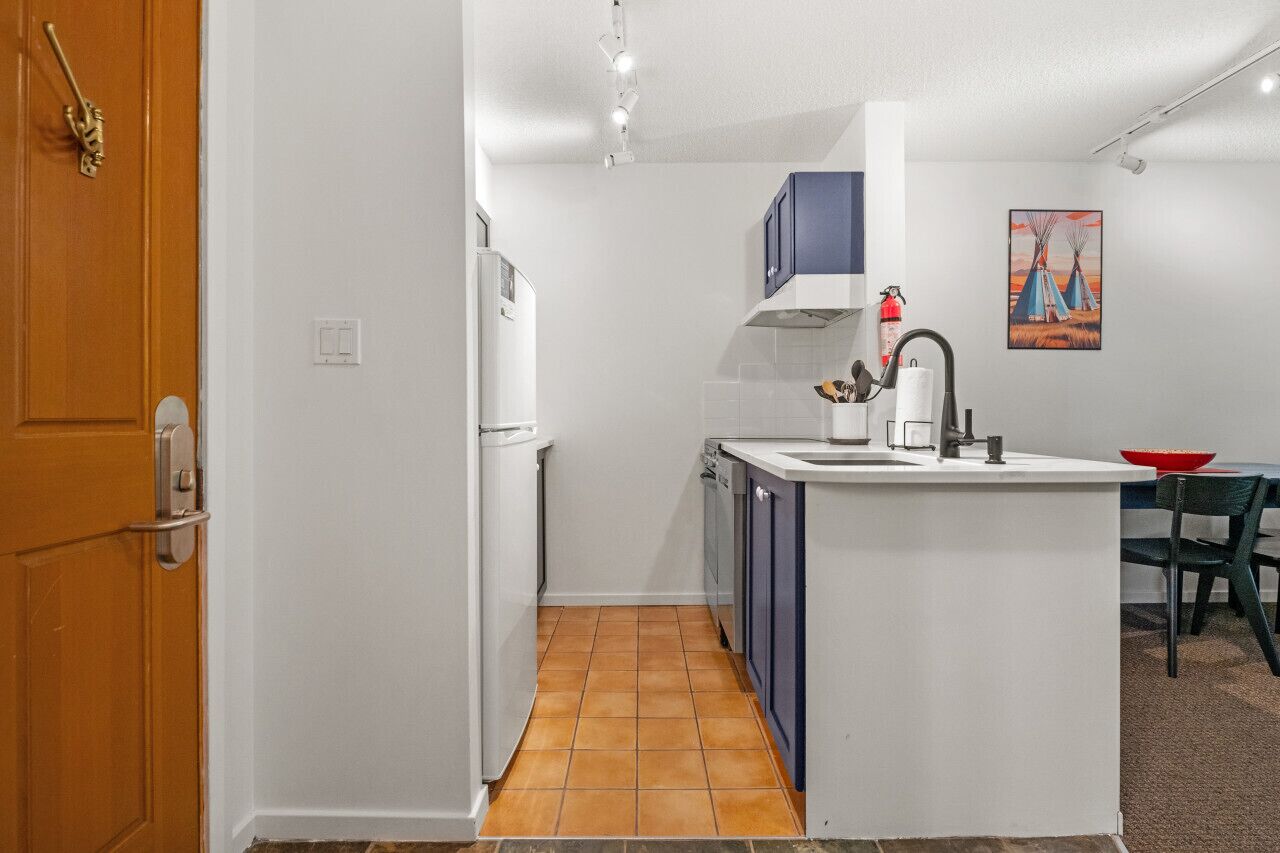 A compact kitchen with a white countertop, a sink, and a stove. There is a wooden door on the left and a dining area with a table and chairs on the right.