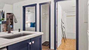 A modern kitchen with dark blue cabinets, a white countertop, and a tiled floor. There is a visible door leading to a bathroom with a shower and a towel rack.