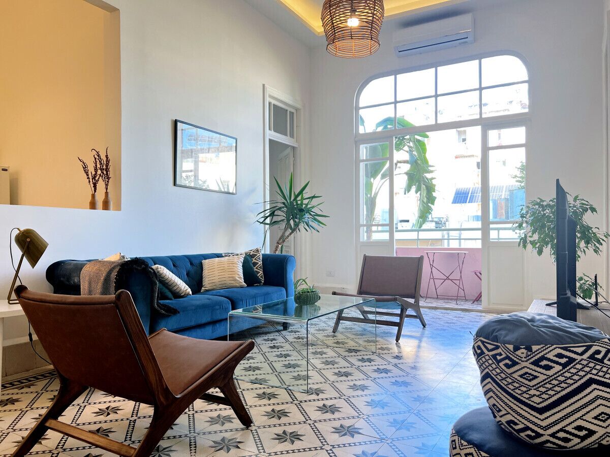 A spacious living room with a blue sofa, a brown armchair, and a patterned coffee table. There is a large window with a view of buildings and a plant in the corner.