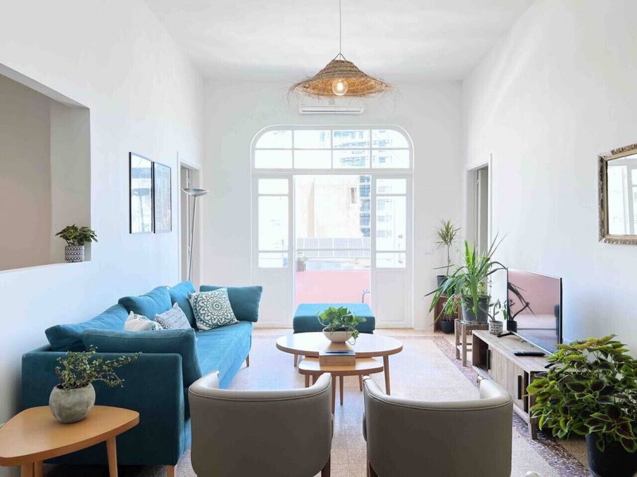 A modern living room with a blue sofa, a round wooden table, and a TV. There are potted plants and a large window providing natural light.