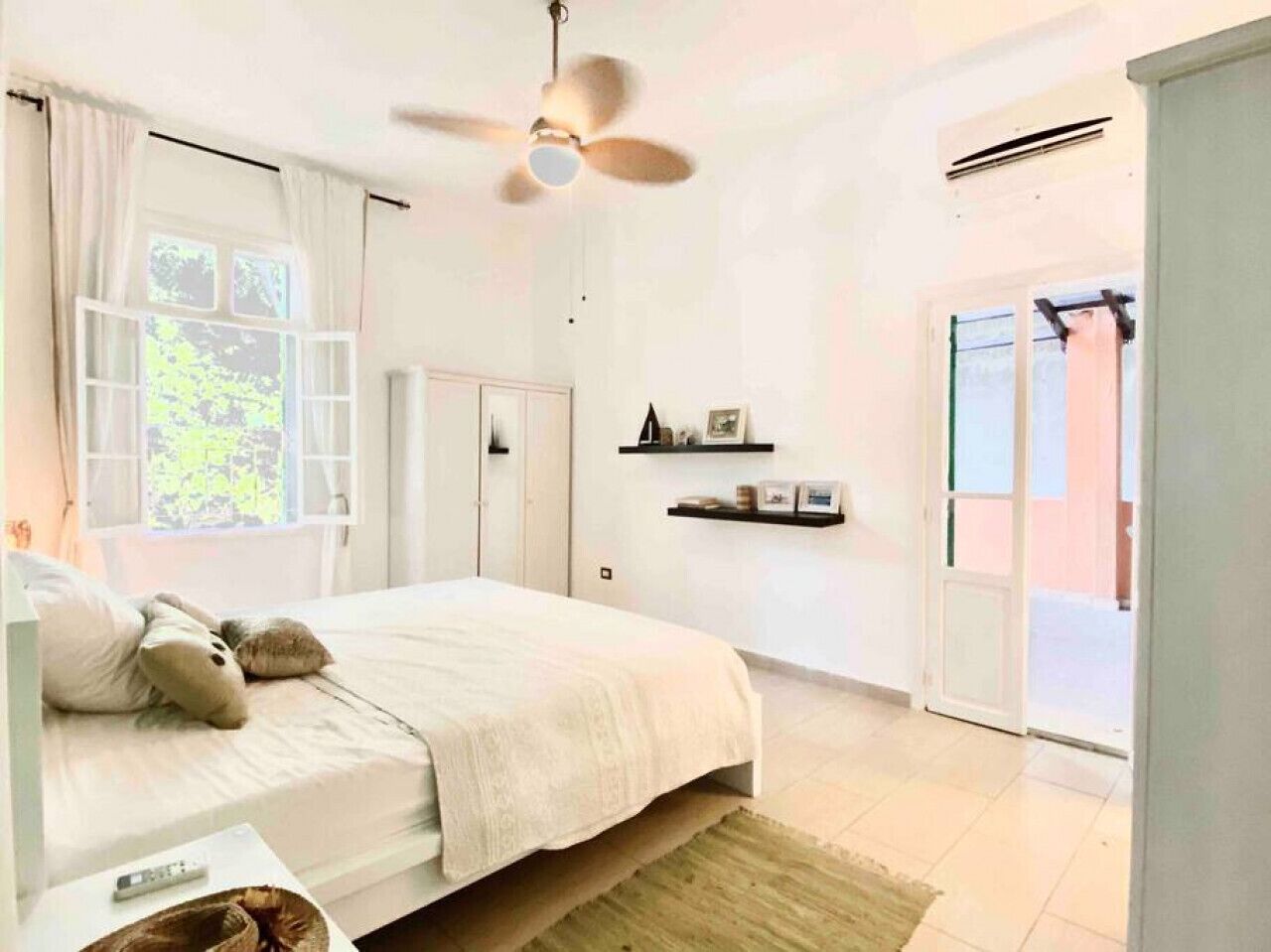 A bedroom with a bed, a ceiling fan, a window with curtains, a door, and a wall-mounted shelf.