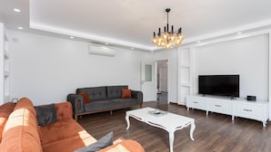 A modern living room with a grey sofa, orange armchairs, a white coffee table, a TV, and a chandelier. - Villa Capital - Five Bedroom Villa, Sleeps 10 (Dalyan)