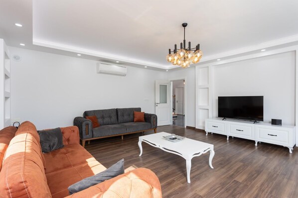 Een moderne woonkamer met een grijze bank, oranje fauteuils, een witte salontafel, een televisie en een kroonluchter.