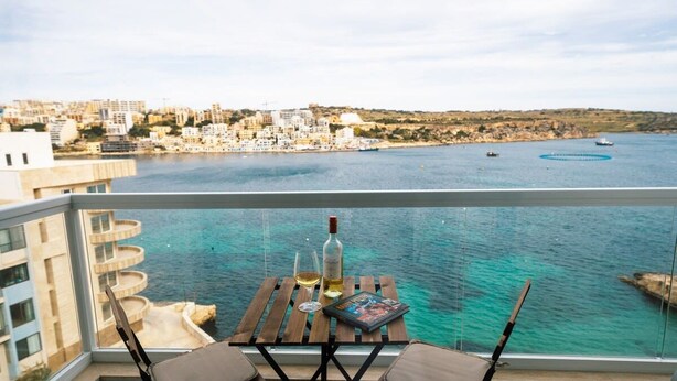 En balkon med et træbord, en vinflaske og et magasin. Udsigten omfatter en kystby og havet.