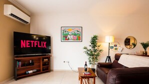 A living room with a TV displaying 'NETFLIX', a framed painting, a lamp, a plant, and a brown leather couch. - A pretty holiday home in Malta’s fishing village (Marsaxlokk)
