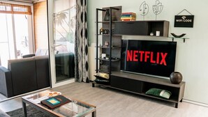 Un salon avec une grande télévision diffusant du contenu « NETFLIX », une table basse en verre, un canapé et une bibliothèque agrémentée d’objets décoratifs.
