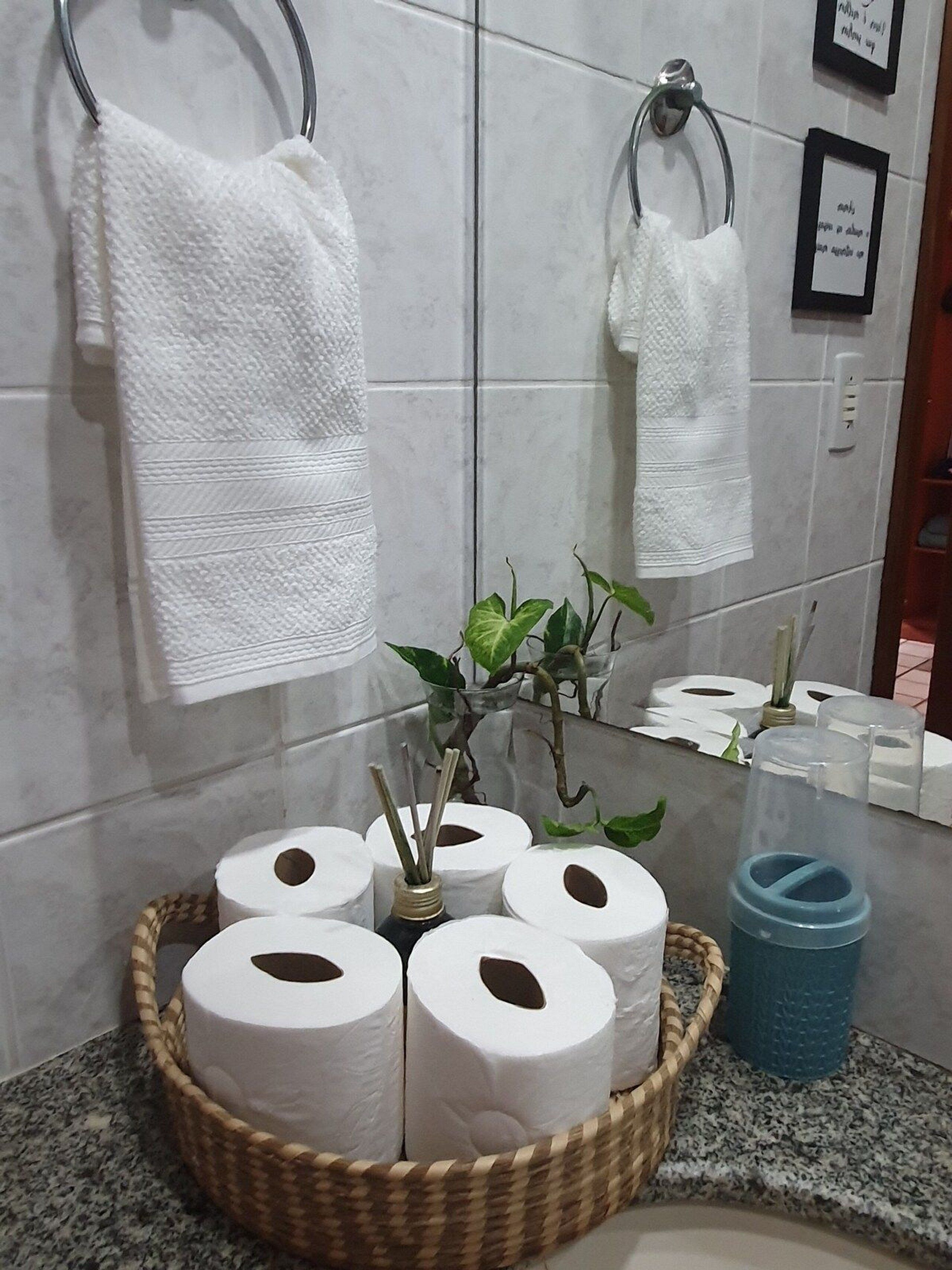 Een badkamer met een betegelde muur, twee witte handdoeken aan metalen houders, een rieten mand met toiletpapierrollen, een blauwe container en een kleine pot met een plantje.
