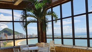 Une terrasse avec une table et des chaises, une grande plante en pot, et une vue sur une plage et le paysage urbain, le tout visible à travers de grandes fenêtres.