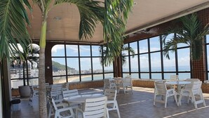 Un espace patio intérieur aménagé avec des tables et des chaises, de grandes fenêtres offrant une vue sur la plage et la ville, ainsi que des palmiers.