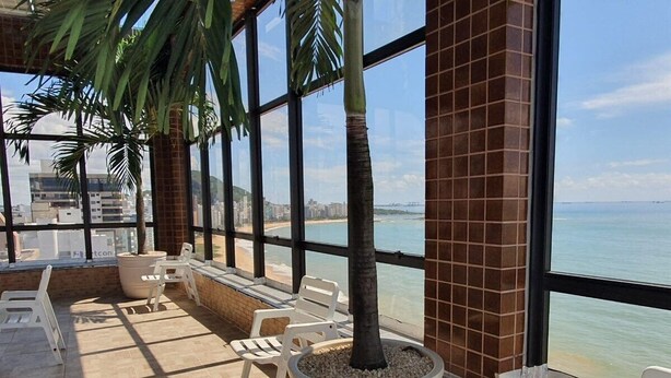 Un espace de détente intérieur avec vue sur la plage, agrémenté de grandes fenêtres, de palmiers en pot et de chaises en plastique blanches.