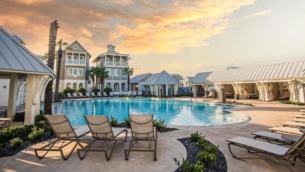 A resort pool area with lounge chairs, a large pool, and resort buildings in the background.