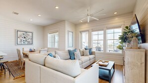 A spacious living room with a large beige sectional sofa, a coffee table, and a TV on a wooden cabinet. There are multiple windows providing natural light and a ceiling fan.