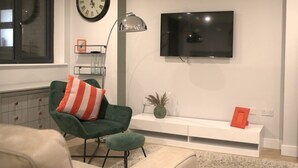 A modern living room with a green armchair, a beige sofa, a large wall clock, a flat-screen TV, and a white console table with decorative items. - Market Eden, King Deluxe suite (Northamptonshire)