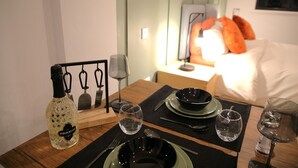 A neatly set dining table with a bottle of Prosecco, black plates, and glasses, in a room with a bed and pillows in the background. - Market Eden, King Deluxe suite (Northamptonshire)