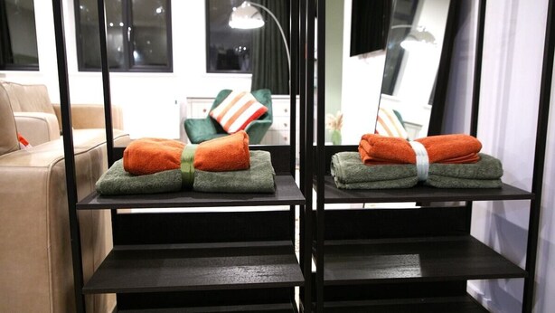 A hotel room with a sofa, a green chair, and a black shelf with folded towels. - Market Eden, King Deluxe suite (Northamptonshire)