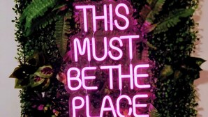 Un insegna al neon con la scritta 'THIS MUST BE THE PLACE' circondata da vegetazione.