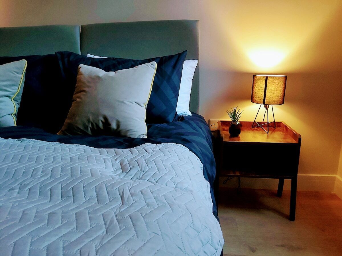 Una cama bien hecha con cubrecama acolchado, una mesita de noche con lámpara y una pequeña planta en maceta.