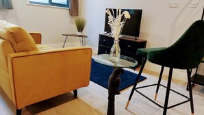 Ein Wohnzimmer mit einem gelben Sofa, einem grünen Sessel, einem Glastisch, einem Fernseher und einer kleinen Pflanze.