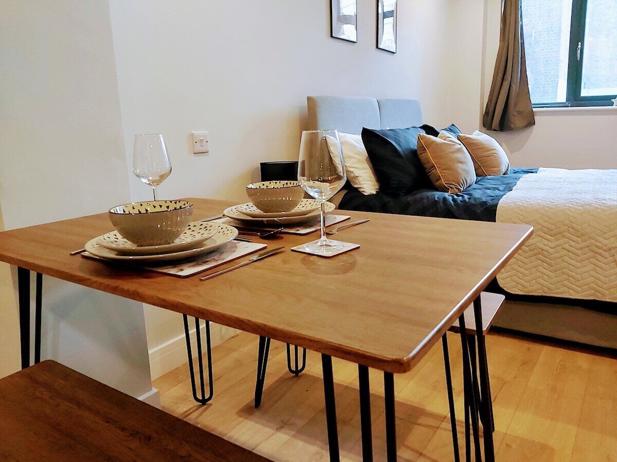 A neatly arranged dining table with plates, bowls, and wine glasses, a bed with pillows, and a window with curtains in a room with wooden flooring.