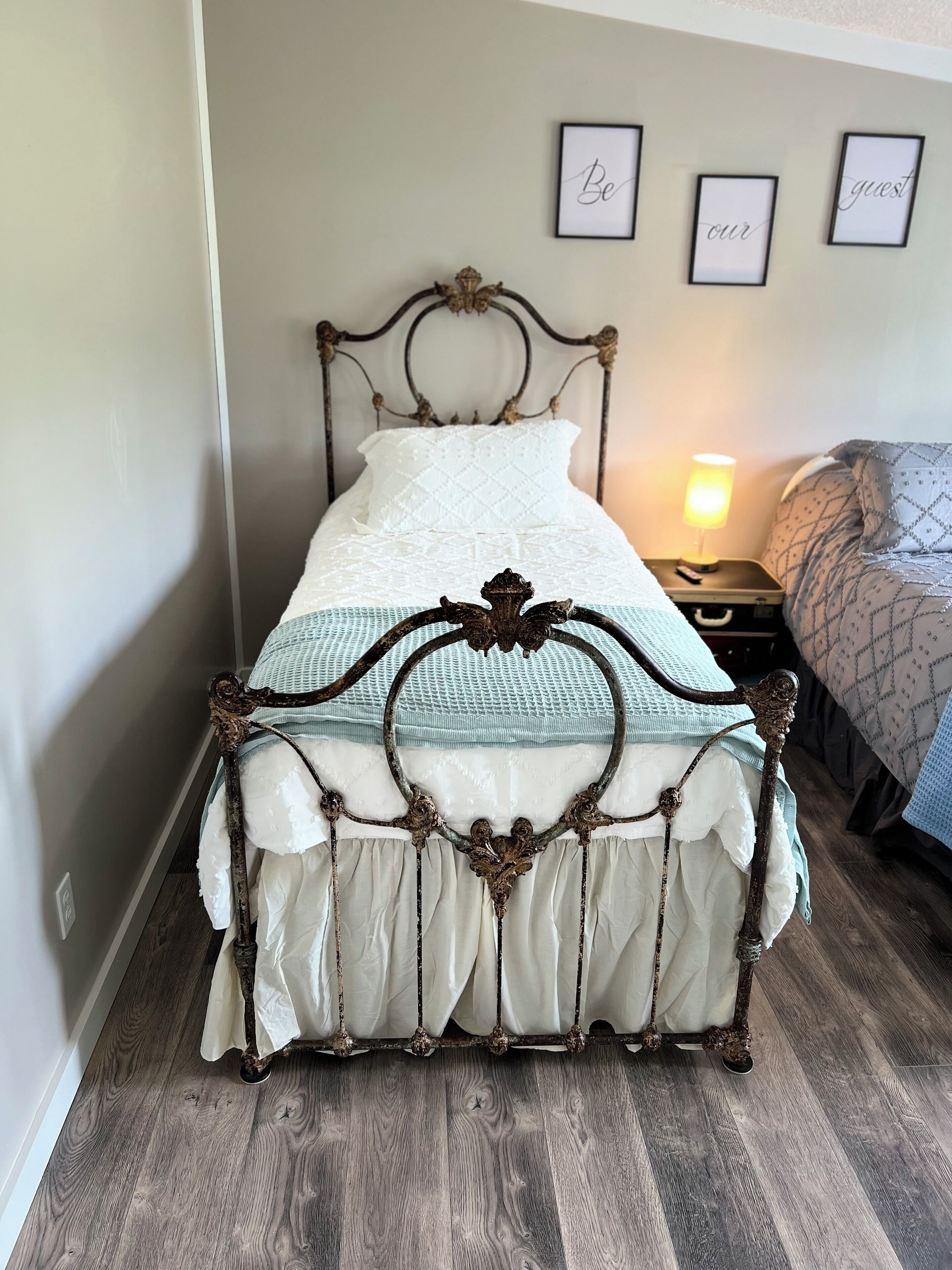 Een bed met een antiek metalen frame, een witte sprei en een bedlampje.