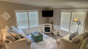 A living room with two beige sofas, a green coffee table, a fireplace, and a flat-screen TV mounted above it. - Cozy Sunset Cottage on the family farm near Cave Run Lake and Morehead State  (Morehead)