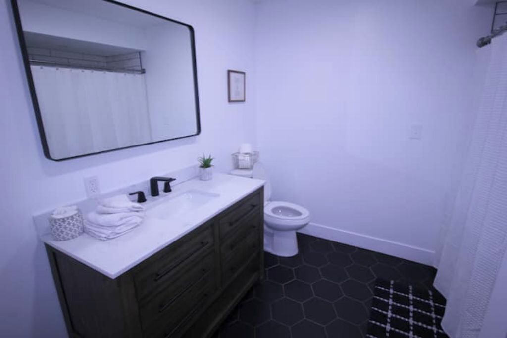 Un baño con un lavamanos blanco, mueble de tocador de madera oscura, inodoro blanco y suelo a cuadros blanco y negro.