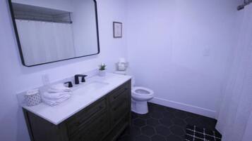 Un baño con un lavamanos blanco, mueble de tocador de madera oscura, inodoro blanco y suelo a cuadros blanco y negro.