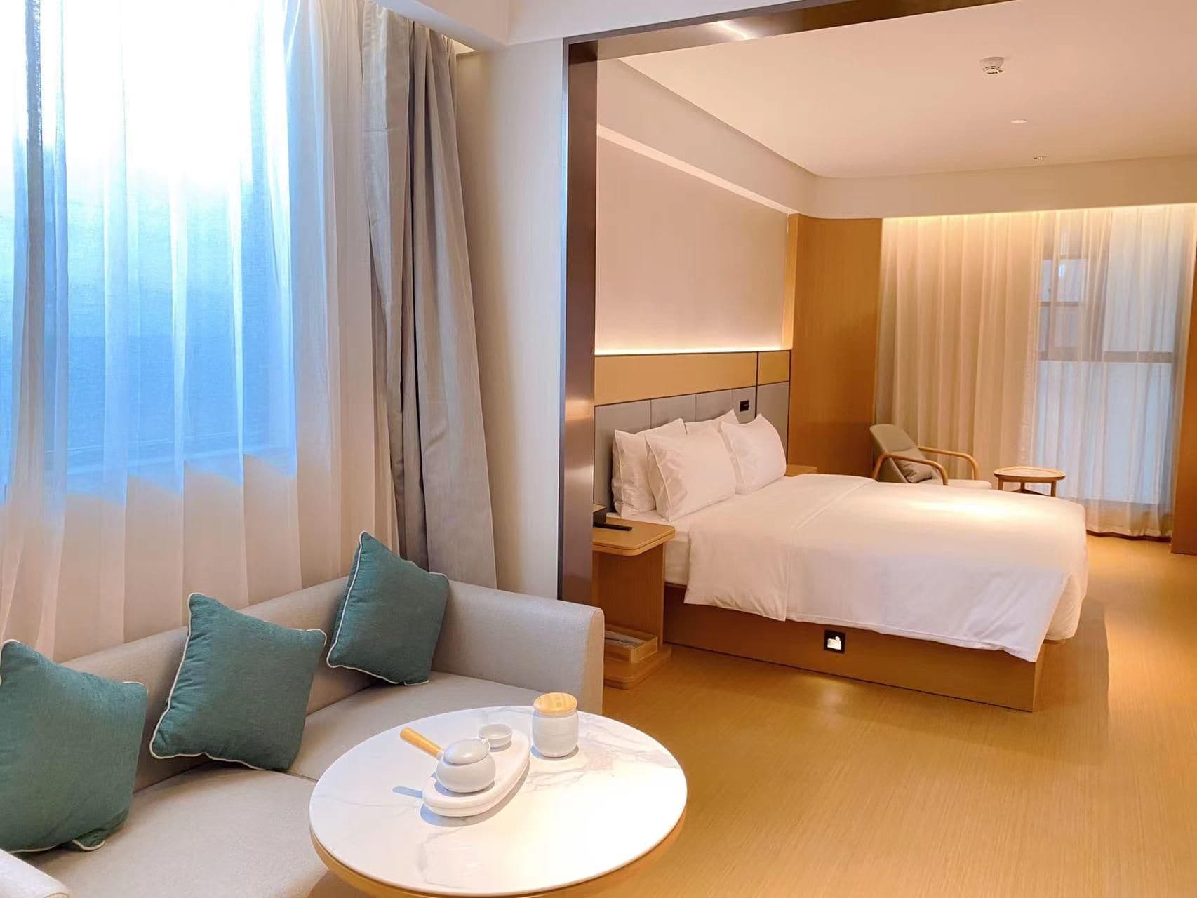 A hotel room with a bed, a sofa, a bedside table with a tea set, and a view through sheer curtains.