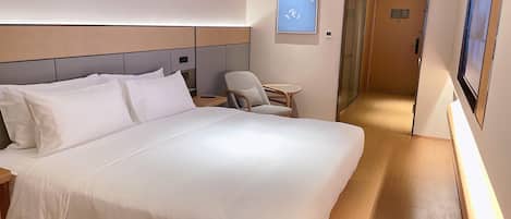 A hotel room with a large bed, a bedside table, a chair, a small table, and a wall-mounted TV.