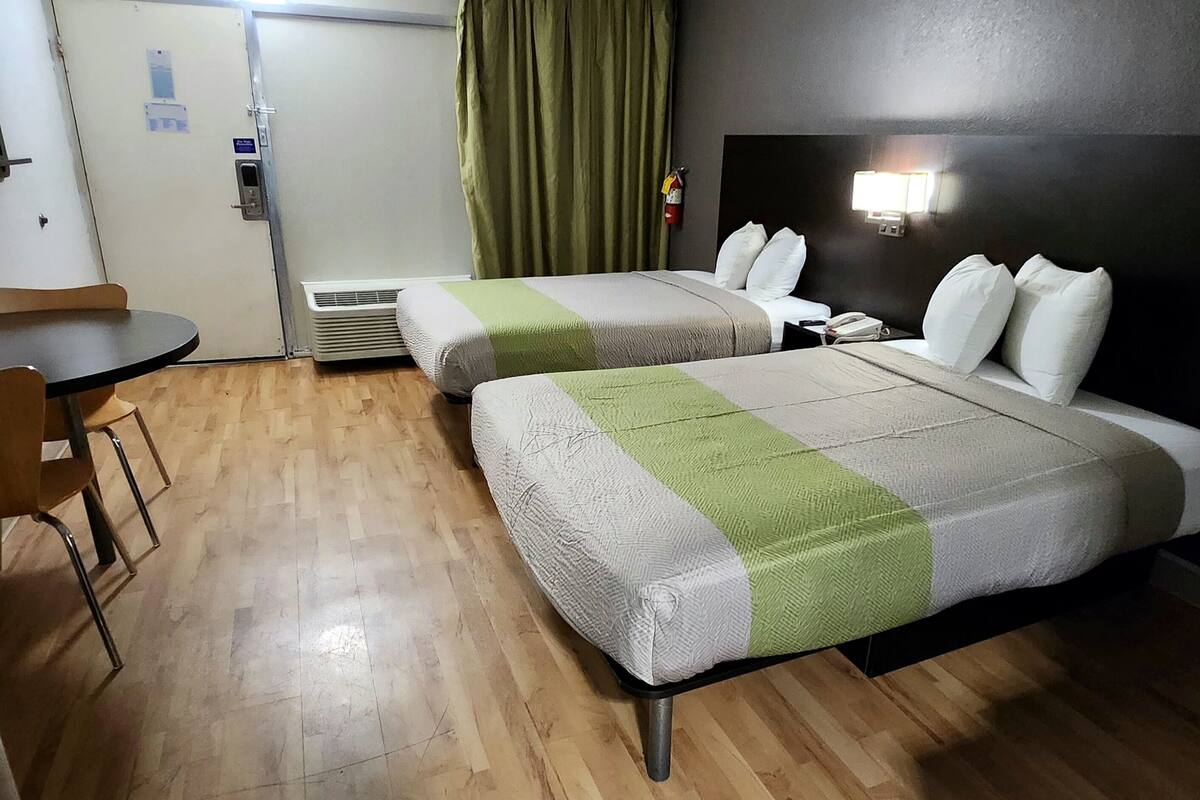 A hotel room with two beds, a small table, a chair, a heater, a door, and a wall-mounted light fixture.