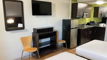 A compact hotel room with a bed, a TV, a small kitchenette, and a chair.