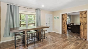 A kitchen with a wooden table and bar stools, a window with curtains, and a door leading to another room.