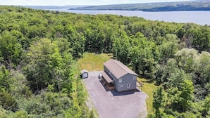 A two-story house with a gravel driveway and a small shed, surrounded by dense forest and a lake in the background.
