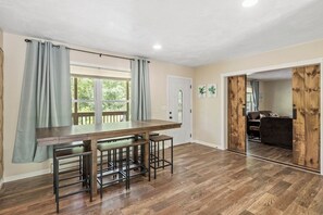 Quarto com mesa de jantar de madeira, banquetas, janela com cortinas, porta e vista para outro quarto com sofá e parede de madeira.