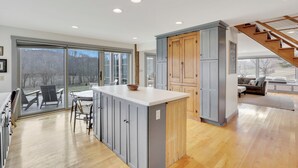 A modern kitchen with a large island, sliding glass doors leading to a deck, and a staircase to a loft area. - Hillside haven on expansive grounds, heated pool, firepit, bocce court and more! (Logan)
