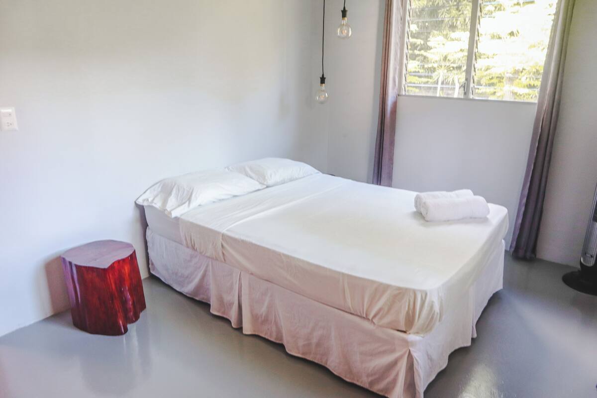 Uma cama bem arrumada com colchão e travesseiros brancos, um banquinho vermelho pequeno e uma janela com cortinas.
