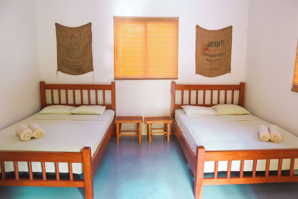 Quarto com duas camas de madeira, uma mesa pequena e janela com persianas.