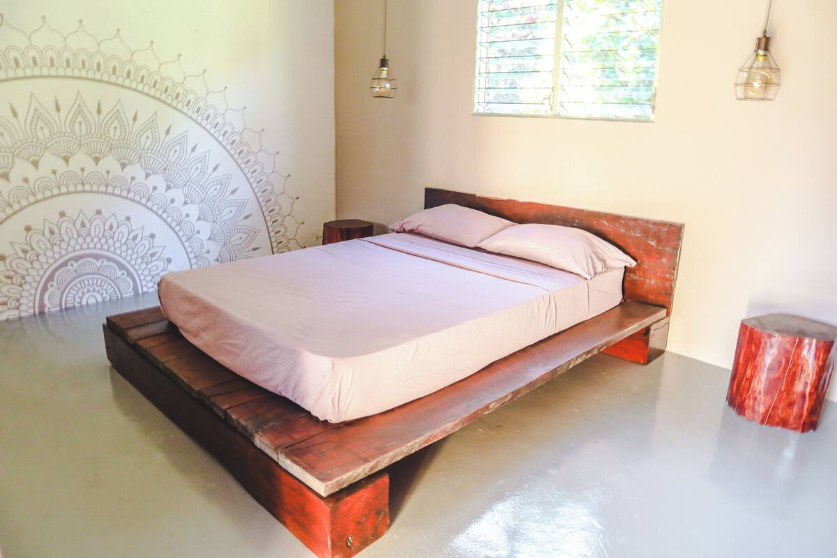 Uma cama com estrutura de madeira e colchão, um banquinho de madeira pequeno e uma parede com padrão decorativo.