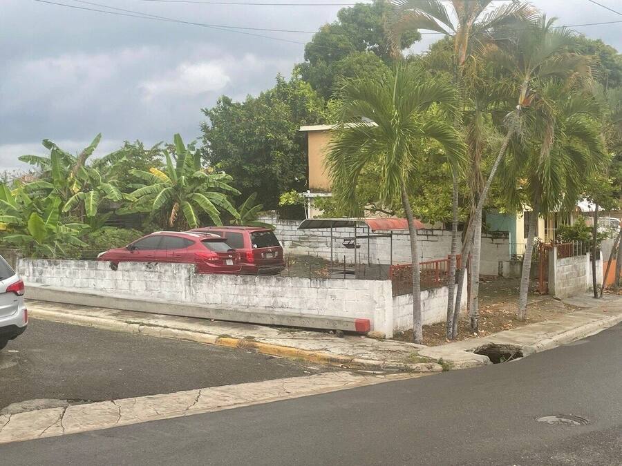 A street view with a white wall, a red car parked on the side, and a building with a red roof in the background.