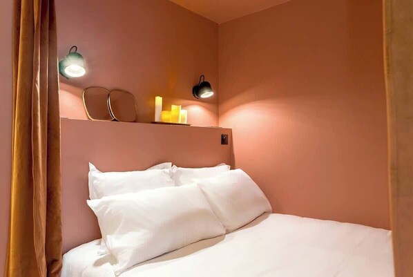 Cama com roupa de cama branca, prateleira com objetos decorativos e iluminação embutida na parede.