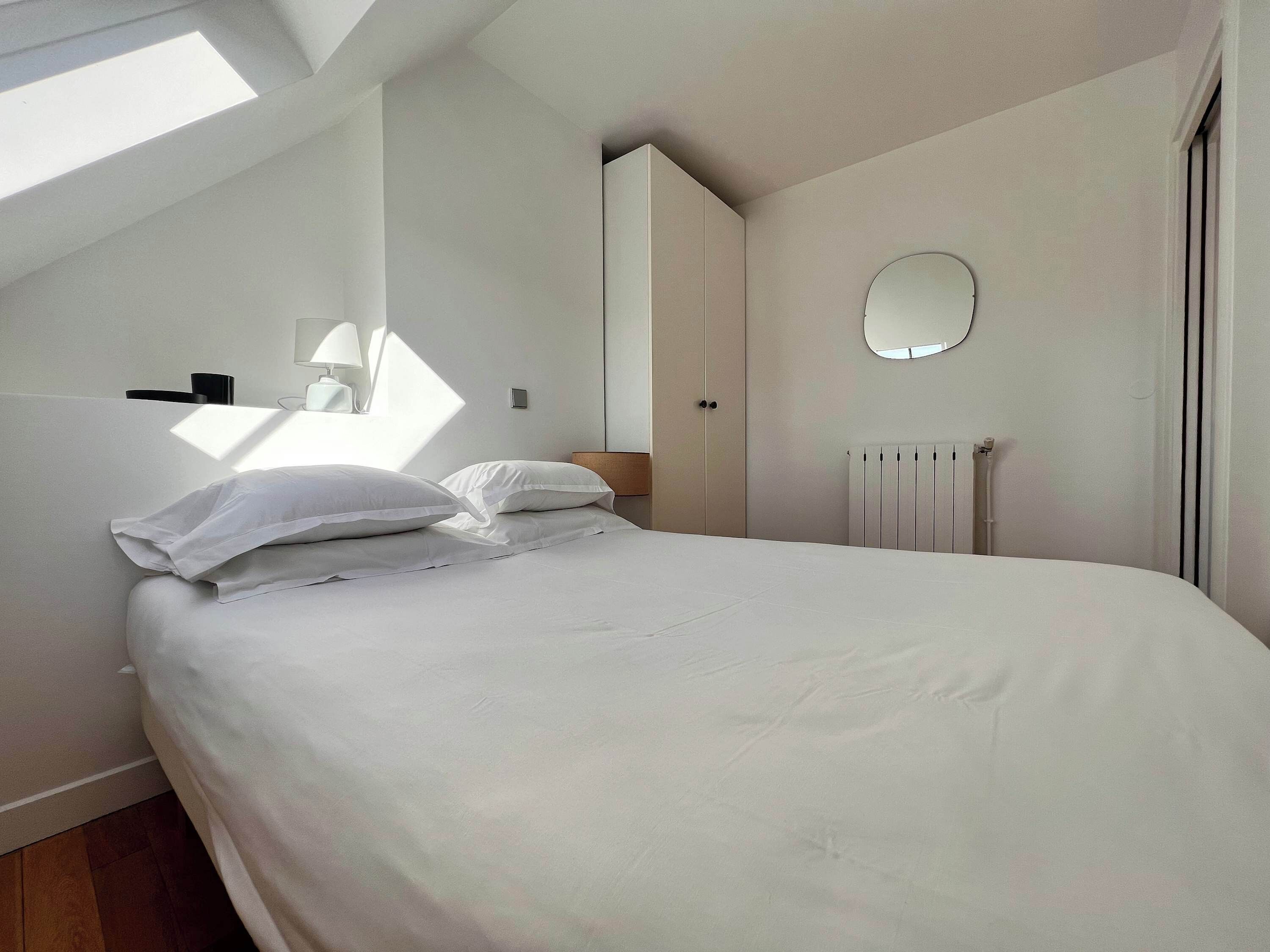 A bedroom with a bed, a bedside table with a lamp, a wardrobe, a radiator, and a skylight.
