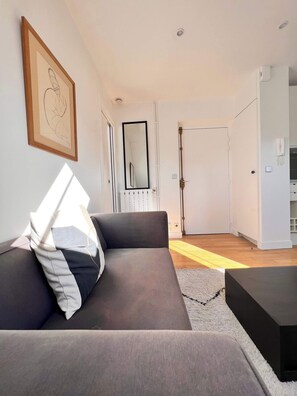 A modern living room with a grey sofa, a black coffee table, a framed artwork on the wall, a large mirror, and a door leading to another room.