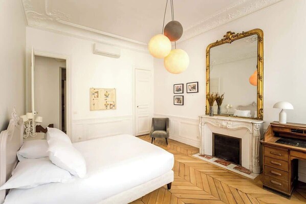Un dormitorio con una cama grande, piso de madera, chimenea, espejo y escritorio.
