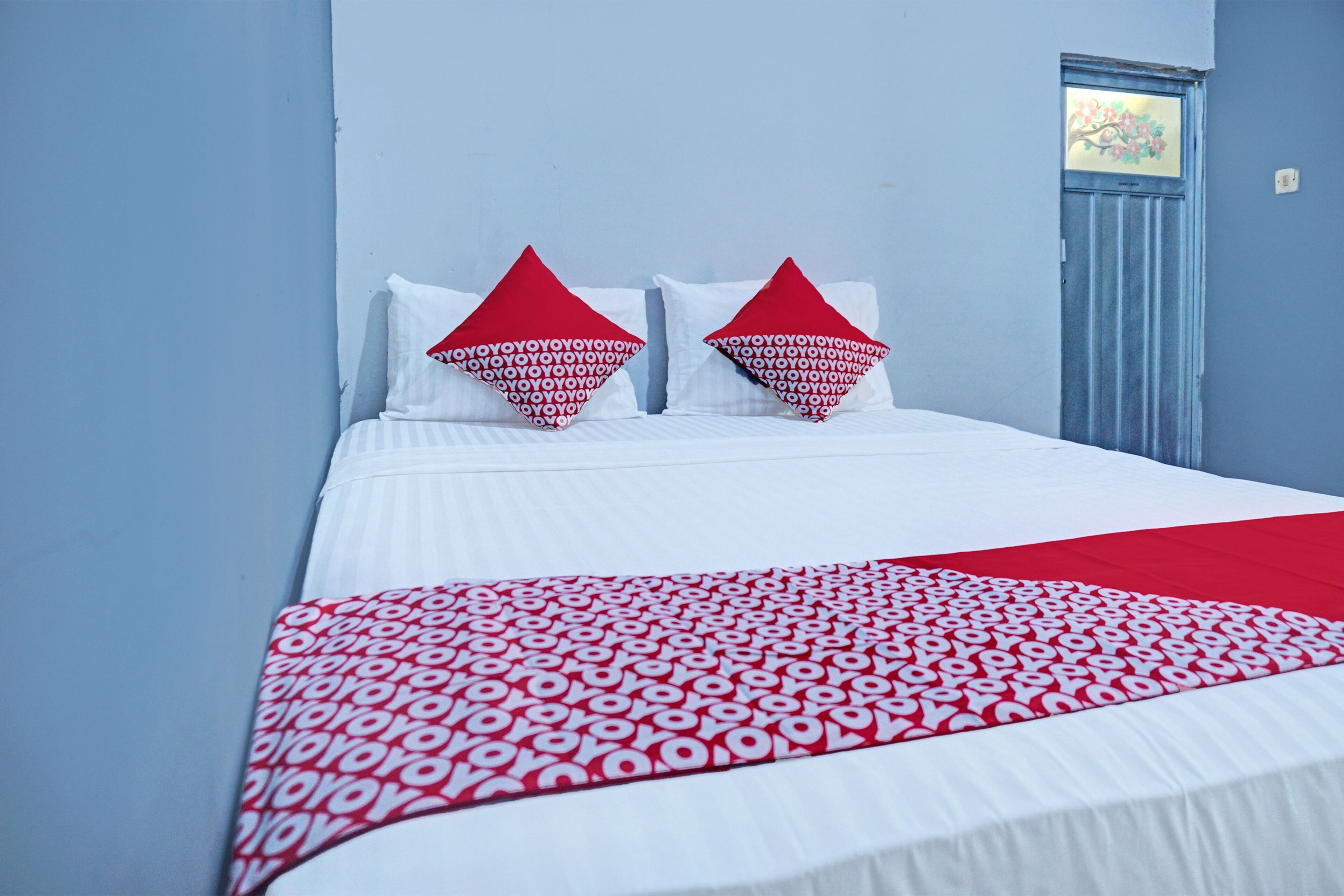 A bed with white bedding and red pillows, a red and white patterned throw blanket, and a blue door with a floral design.