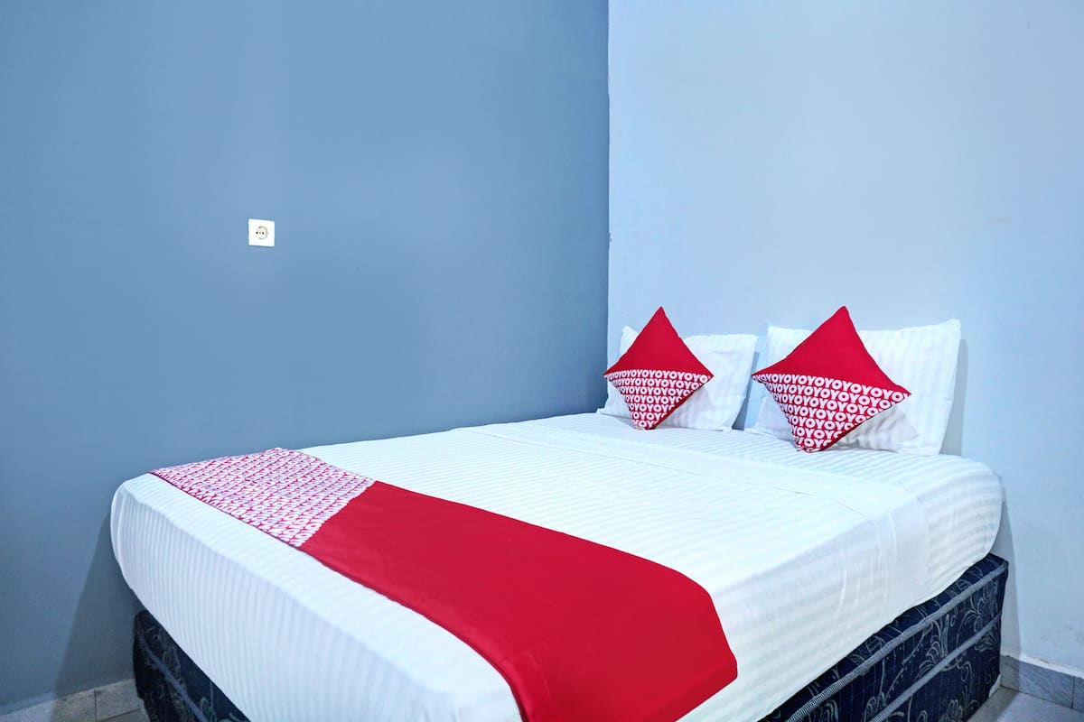 Un lit bien fait, recouvert d’une couvre-lit blanche et rouge, avec deux oreillers rouges et une tête de lit sombre, le tout adossé à un mur bleu.