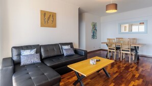 A modern living room with a black leather sofa, wooden coffee table, and a dining area with wooden chairs and a table. - Estrela Azul 5AR (Caniço)