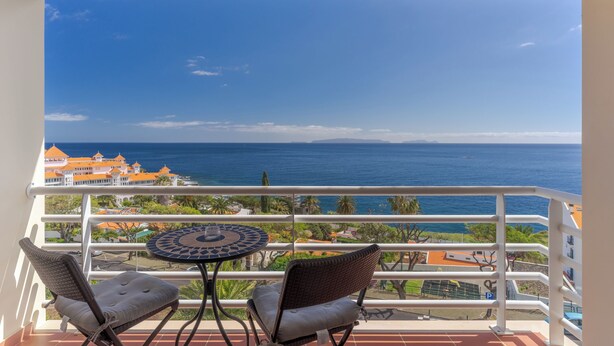 A balcony with two wicker chairs and a small round table, overlooking a coastal view with buildings and the sea. - Estrela Azul 5AR (Caniço)