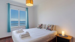 A neatly made bed with folded towels, a bedside table with a candle, and a window with curtains. - Estrela Azul 5AR (Caniço)