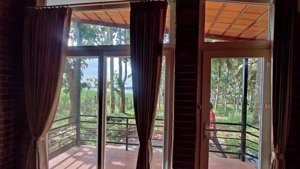A room with a large sliding glass door leading to an outdoor area with a metal railing and a view of greenery and a body of water. - Kabini Earthern Cottages Lakeside Farmstay
 (Holehundi)