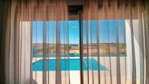 A view through sheer curtains of a swimming pool and a landscape with trees and open fields. - Hôtel Debrazza (Ouled Tayeb)